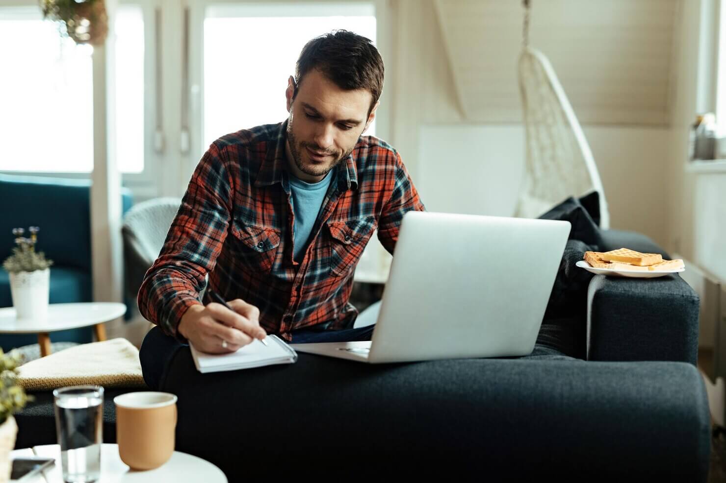Smiling man writing notes while using computer in the living room. Writing out key eCommerce features to build