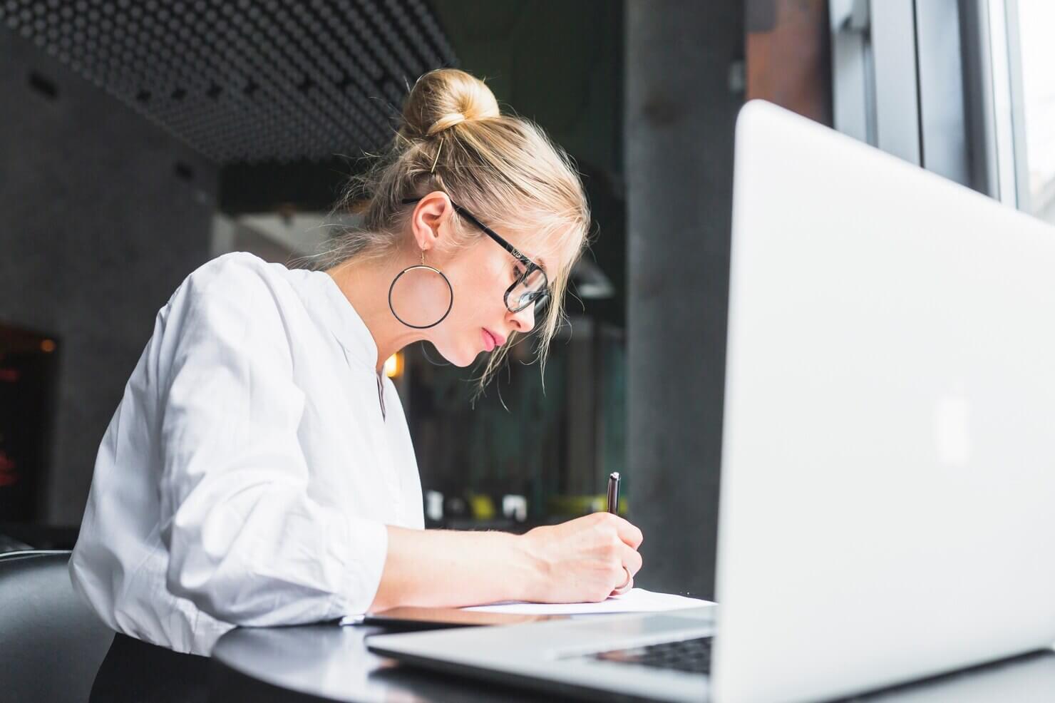 Woman writing on document with laptop on desk. She is writing about automated WordPress maintenance options